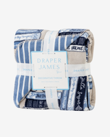 A folded decorative throw blanket with a blue, white, and beige patterned print featuring book spines and script text, wrapped in white paper bands with the brand name Draper James visible.