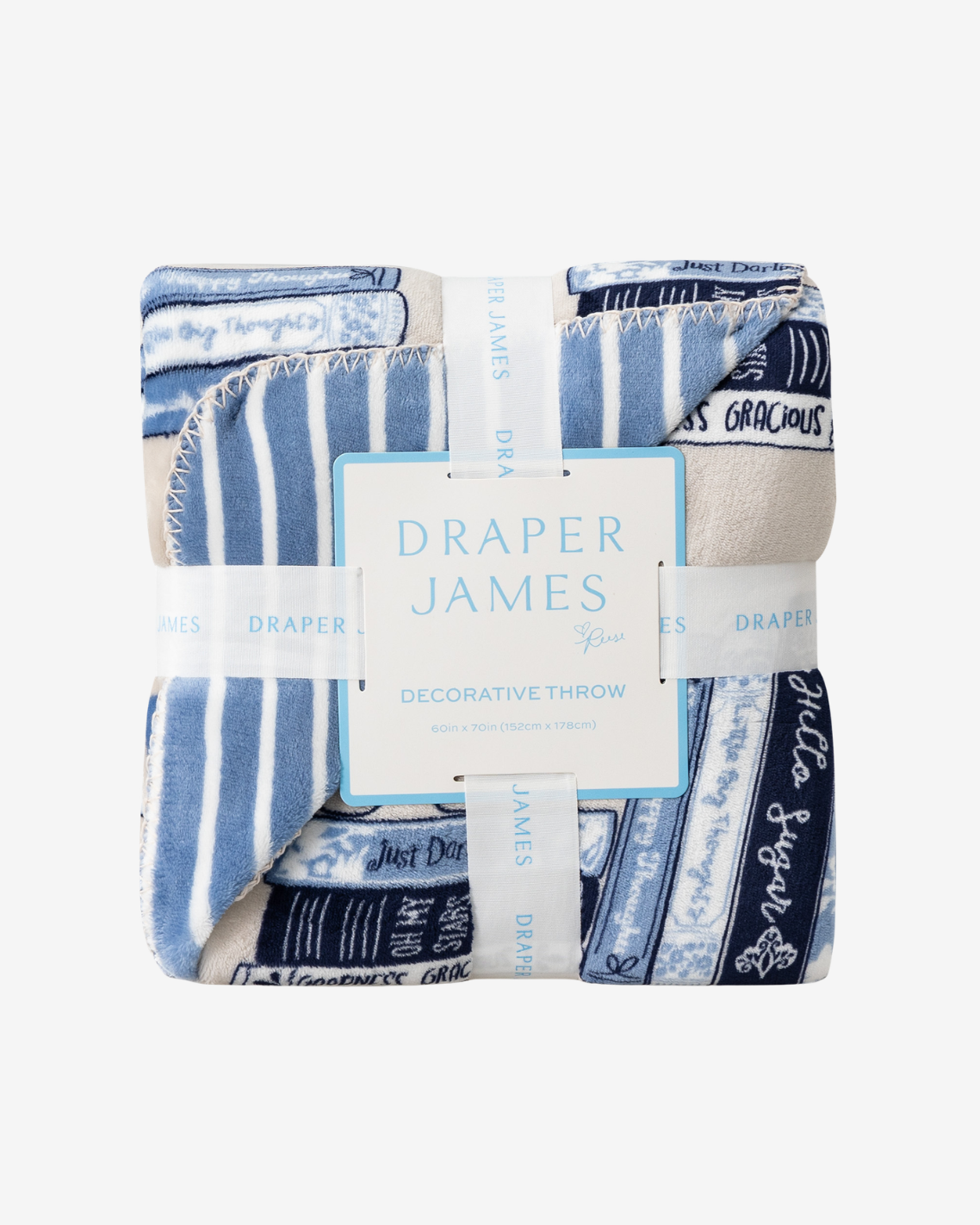 A folded decorative throw blanket by Draper James, featuring a blue and white striped pattern with navy text bands and a central white paper label, photographed on a light gray background.