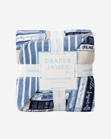 A folded decorative throw blanket by Draper James, featuring a blue and white striped pattern with navy text bands and a central white paper label, photographed on a light gray background.