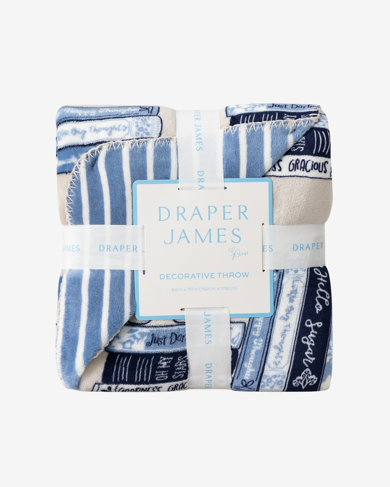 A folded decorative throw blanket by Draper James, featuring a blue and white striped pattern with navy text bands and a central white paper label, photographed on a light gray background.