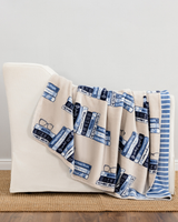 A cream throw blanket with a blue and navy book-and-texture print draped over a white armchair in a styled room setting.