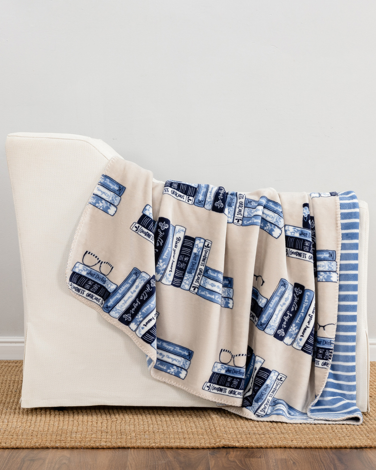 A cream throw blanket with a blue and navy book-and-texture print draped over a white armchair in a styled room setting.