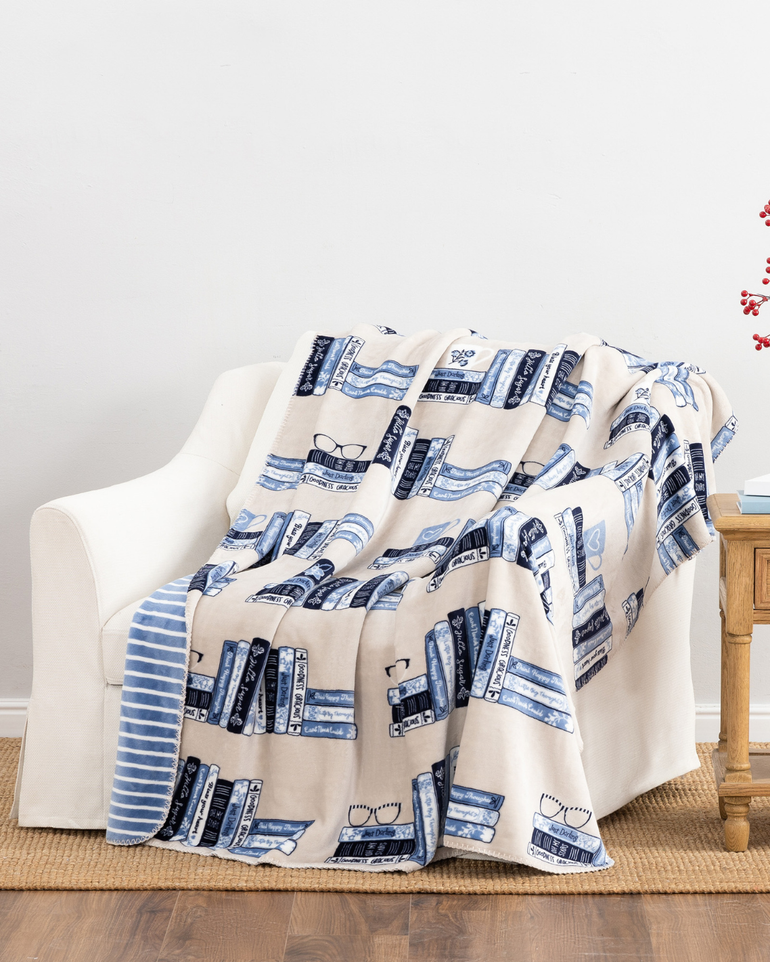 A cream-colored throw blanket with a blue and navy book-and-glasses pattern, draped over a white armchair in a living room setting.