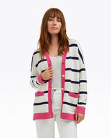 A woman models the Draper James Cassadee Cardigan, a V-neck cotton-wool sweater with pink trim and navy stripes, layered over a white top and white pants against a plain white background.
