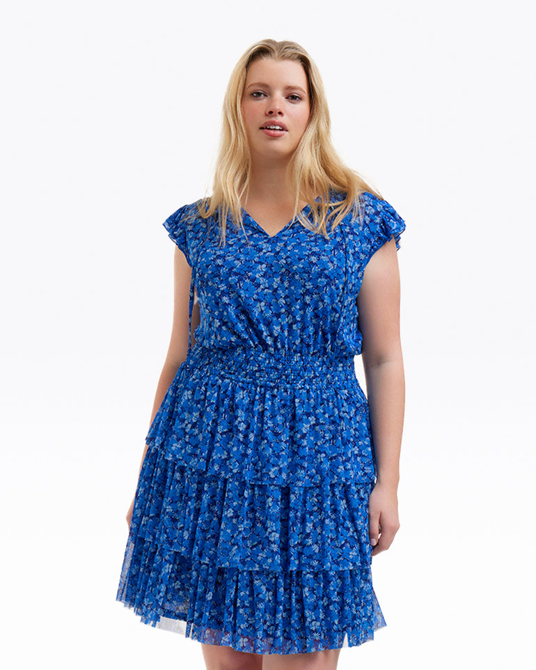 A woman wears the Draper James Alana Tiered Dress in Blue Scattered Ditsy, featuring short sleeves and a ruffled skirt, standing against a plain white background.