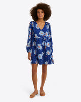 A woman stands facing forward in the Draper James Kellie Babydoll Dress in Printed Dot Jacquard, featuring a blue floral pattern and v-neckline, paired with tan flat sandals.