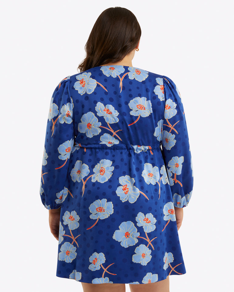 A woman is shown from the back wearing the Draper James Kellie Babydoll Dress in Printed Dot Jacquard, featuring a blue v-neck, long sleeves, and a light blue floral pattern, set against a plain background.