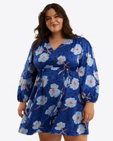 A woman in the Draper James Kellie Babydoll Dress in Printed Dot Jacquard, featuring blue floral print, long sleeves, a v-neck, and gathered waist, stands against a plain white background.