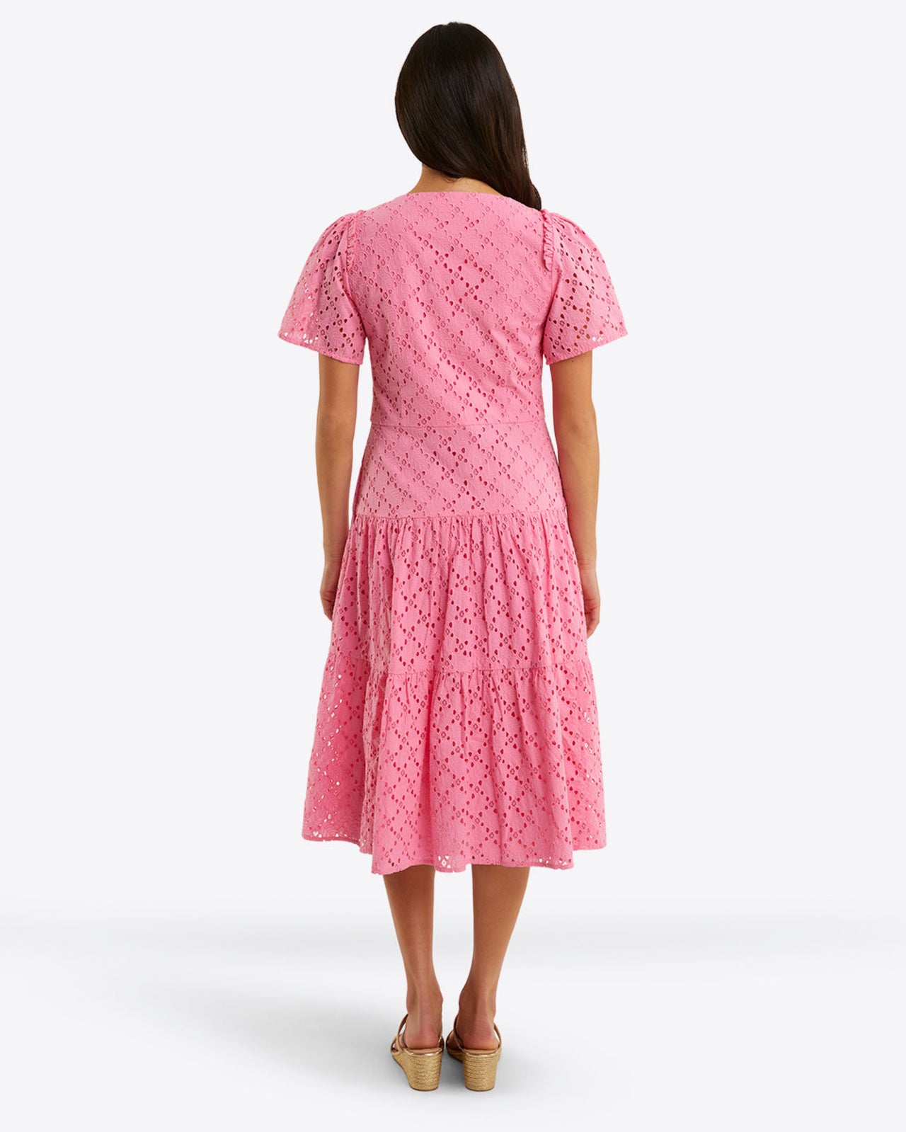 A person stands facing away, wearing the Draper James Caitlyn Midi Dress in pink cotton eyelet, featuring a square neckline, short sleeves, and a tiered skirt.