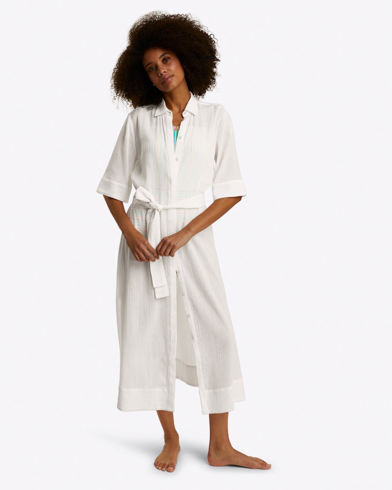 A woman stands barefoot in the Draper James Crinkle Cotton Coverupâ€”a white button-down beach dress with a removable belt and three-quarter sleevesâ€”against a plain background.