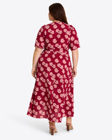 A woman wearing the Draper James Fiona Wrap Midi Dress in Lurex Crepe—a red and pink floral-patterned midi with short sleeves and a high-low hem—stands with her back to a plain background.