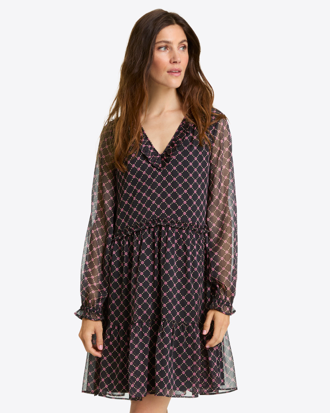 A woman wears the Draper James Lorrie Long-Sleeve Dress in Crinkle Chiffonâ€”a black, V-neck dress with pink and white diamond print, ruffled hem, and long sleevesâ€”standing against a plain background.