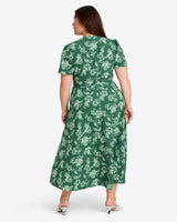 A woman faces away, wearing the Gigi Wrap Dress by Draper Jamesâ€”a green, short-sleeved, ankle-length party dress with a white floral patternâ€”and white-heeled sandals.