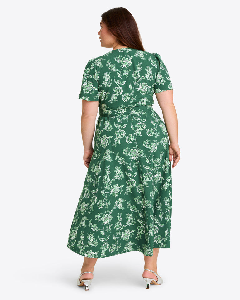 A woman faces away, wearing the Gigi Wrap Dress by Draper Jamesâ€”a green, short-sleeved, ankle-length party dress with a white floral patternâ€”and white-heeled sandals.