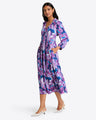 A woman in the bold floral Katie Midi Dress by Draper James stands with one hand in her pocket and the other on her hip, paired with white pointed shoes.
