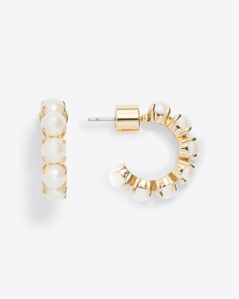 The Draper James Huggie Hoops in Pearl feature stunning white pearls on gold, with one earring facing forward and the other sideways against a crisp white background, adding timeless elegance to any ensemble.