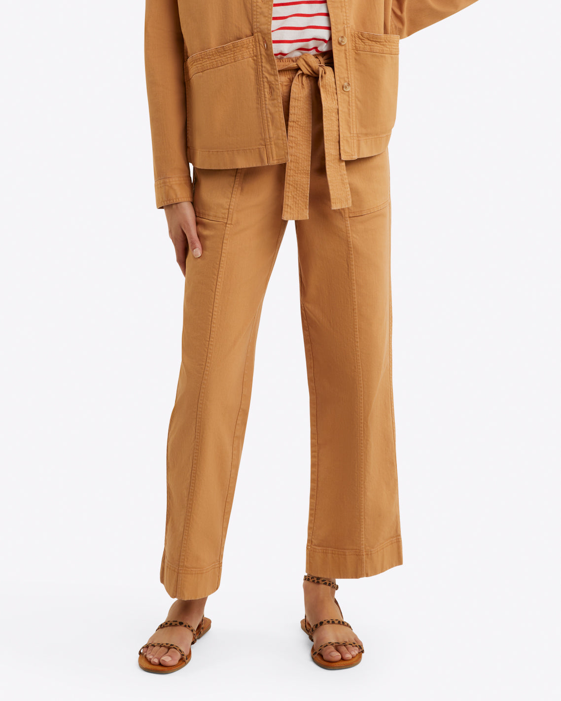 A person is styled in a tan ensemble from Draper James, featuring a belted jacket, relaxed-fit Utility Pant in Cotton Twill, a striped shirt, and sandals on a white background.