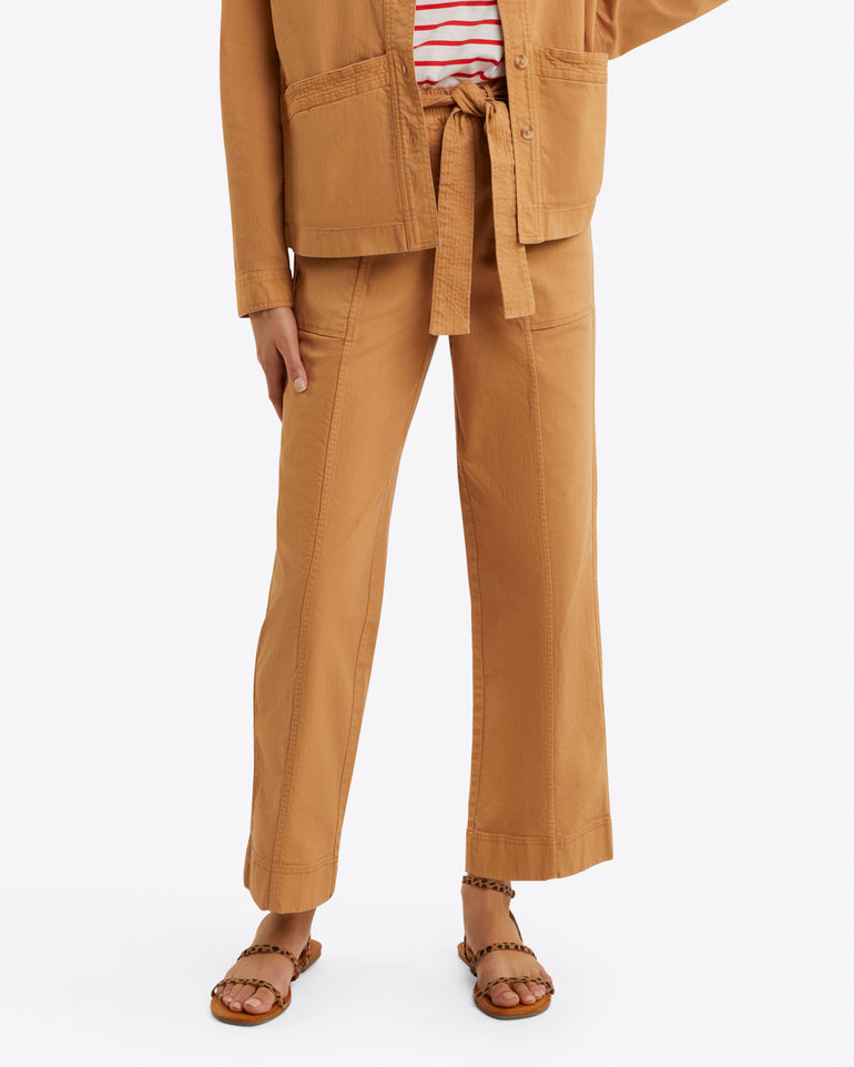 A person is styled in a tan ensemble from Draper James, featuring a belted jacket, relaxed-fit Utility Pant in Cotton Twill, a striped shirt, and sandals on a white background.