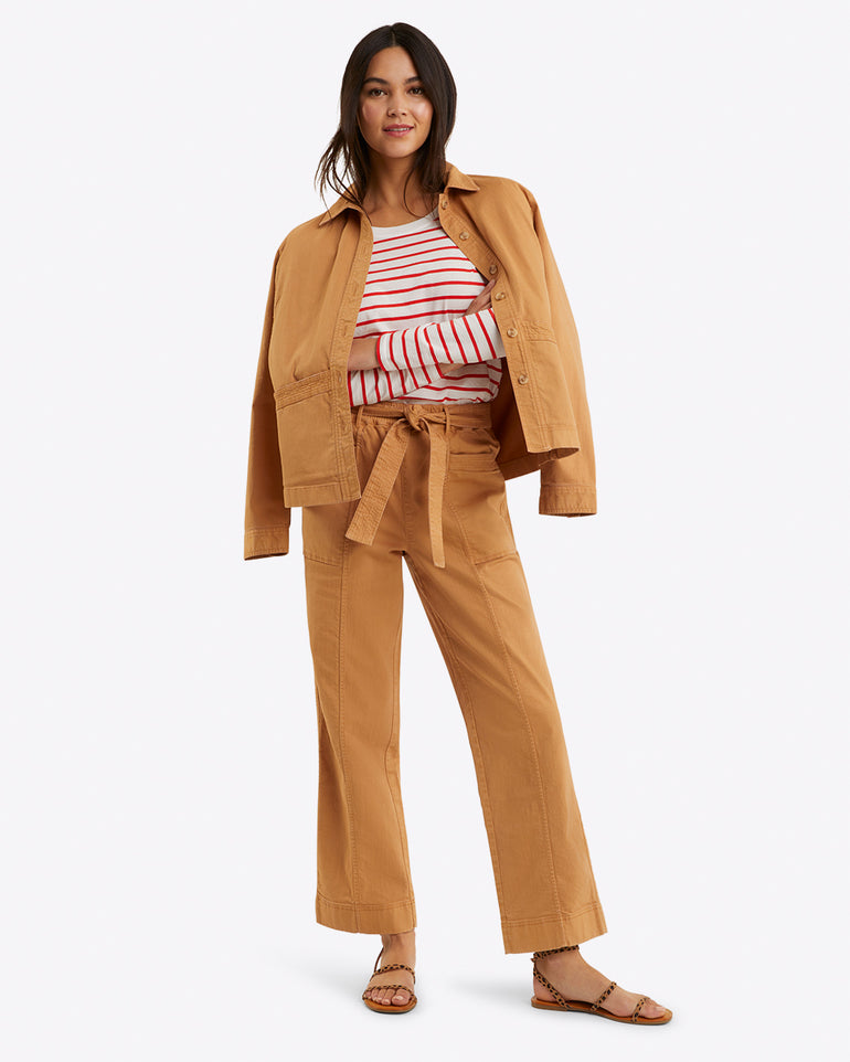 A person models Draper James' Utility Jacket in Cotton Twill with matching pants over a striped shirt, smiling in sandals, perfect for spring layering against a simple backdrop.