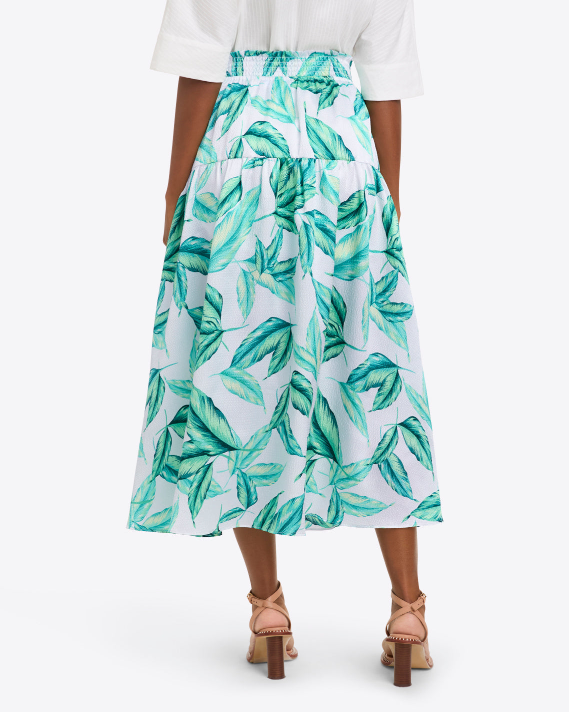 A woman, seen from the back, wears the Draper James Midi Skirt in Hammered Satin featuring a green leaf print and smocked waist, paired with a white top and strappy heeled sandals.