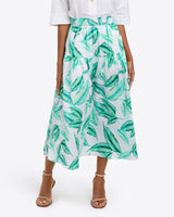 A woman wearing a white top and the Draper James Midi Skirt in Hammered Satin, featuring a green leaf print, smocked waist, and ankle-length light blue fabric, paired with tan heeled sandals.