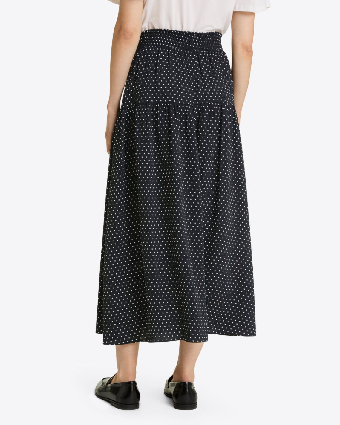 A person is seen from the back wearing the Draper James Midi Skirt in Polka Dot, featuring black fabric with small white spots, paired with a white top and black shoes.