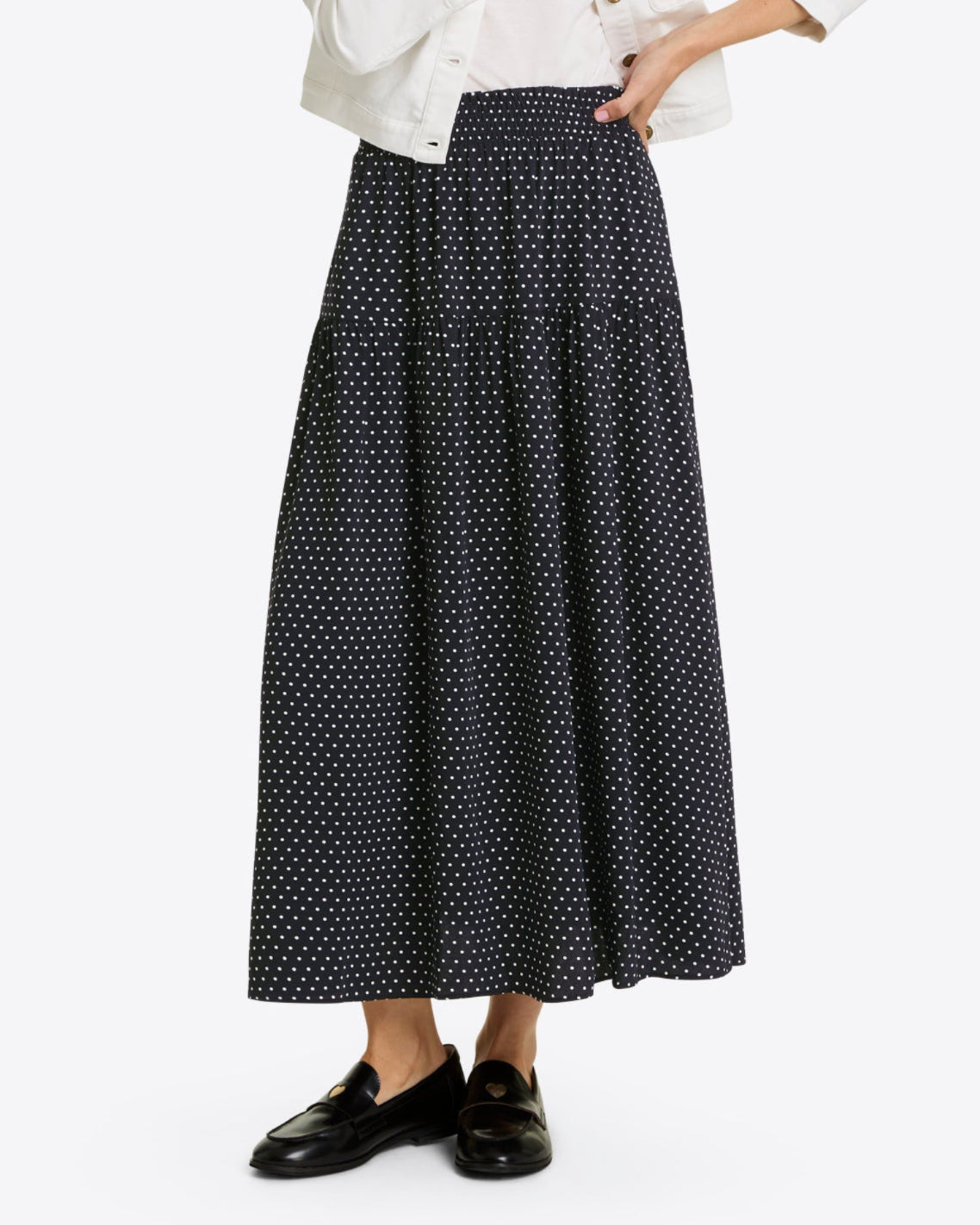 A person wearing the Draper James Midi Skirt in Polka Dot, paired with a white jacket and black loafers, stands against a plain background.