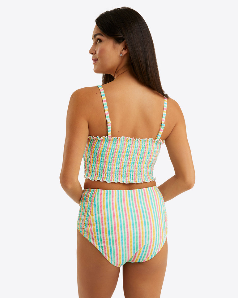 A woman models the Draper James Smocked Bikini Bottom—rainbow-striped and high waisted—paired with a smocked bikini top, facing slightly away from the camera against a plain white background.