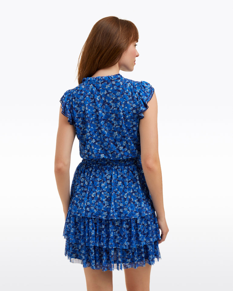 A woman with brown hair is shown from the back wearing the Draper James Alana Tiered Dress in Blue Scattered Ditsy, featuring short sleeves and a tiered skirt, standing against a plain white background.