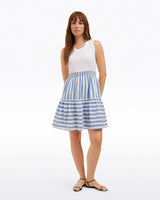 A woman stands against a plain white background wearing the Draper James Arden Mixed Media Shirtdress in Wave Stripeâ€”a sleeveless white top with a blue-and-white striped skirtâ€”paired with black sandals.