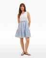 A woman stands against a plain white background wearing the Draper James Arden Mixed Media Shirtdress in Wave Stripeâ€”a sleeveless white top with a blue-and-white striped skirtâ€”paired with black sandals.