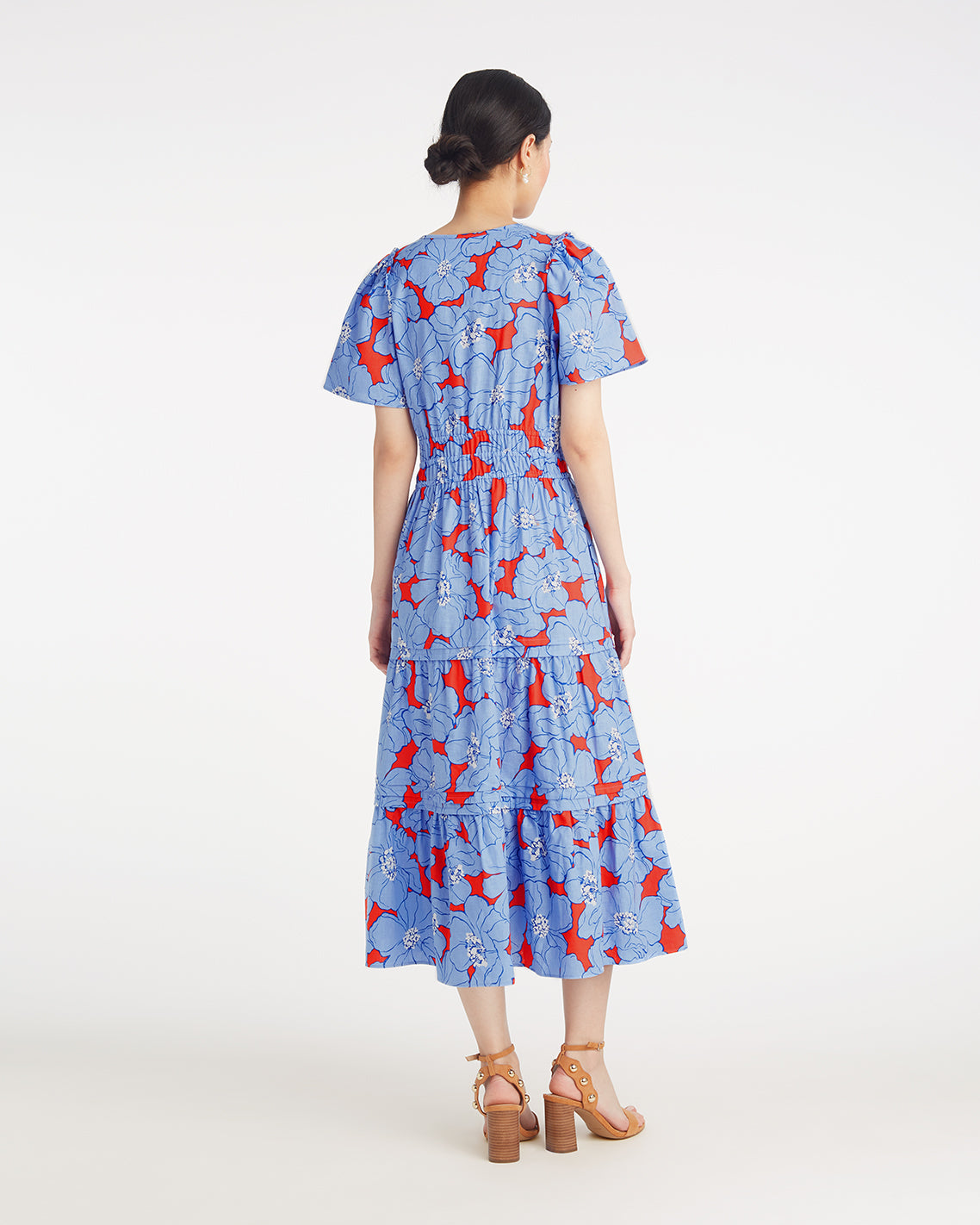 A woman faces away, wearing the Draper James Hollie Midi Dress in Floating Floral—a blue, ankle-length cotton-linen dress with red and white floral patterns—and brown heeled sandals against a plain white background.