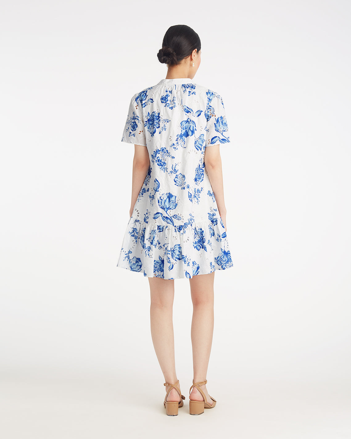 A woman faces away, wearing the Draper James Molly Mini Dress in Magnolia Toile—a white cotton dress with blue floral print and short sleeves, styled with beige sandals for effortless comfort and style.