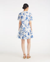 A woman faces away, wearing the Draper James Molly Mini Dress in Magnolia Toile—a white cotton dress with blue floral print and short sleeves, styled with beige sandals for effortless comfort and style.
