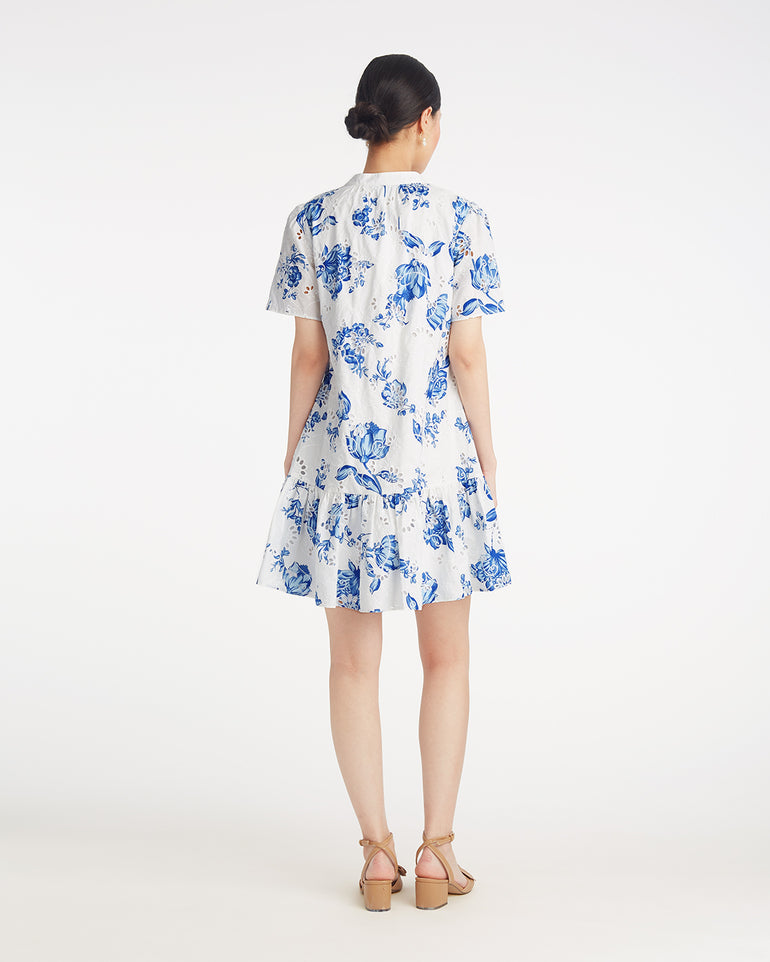 A woman faces away, wearing the Draper James Molly Mini Dress in Magnolia Toile—a white cotton dress with blue floral print and short sleeves, styled with beige sandals for effortless comfort and style.