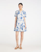 A woman stands against a white background wearing the Draper James Molly Mini Dress in Magnolia Toile, a short-sleeve, knee-length 100% cotton dress with blue magnolia patterns, paired with tan sandals.