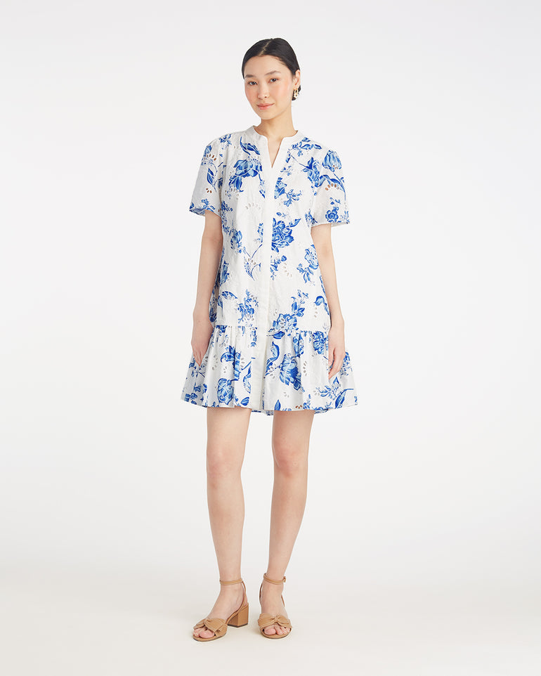 A woman stands against a white background wearing the Draper James Molly Mini Dress in Magnolia Toile, a short-sleeve, knee-length 100% cotton dress with blue magnolia patterns, paired with tan sandals.