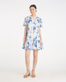 A woman stands against a white background wearing the Draper James Molly Mini Dress in Magnolia Toile, a short-sleeve, knee-length 100% cotton dress with blue magnolia patterns, paired with tan sandals.