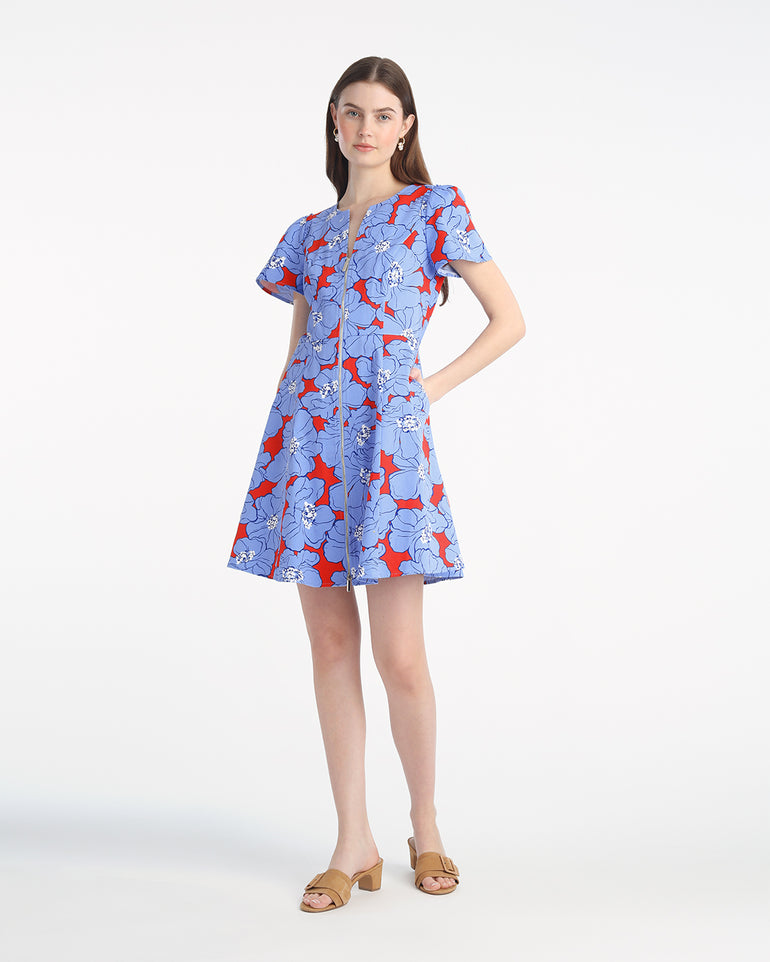 A woman stands against a plain background, wearing the Draper James Mae Love Circle Mini Dress in Floating Floral, featuring blue and red florals, short sleeves, a mini hemline, and tan sandals.