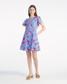 A woman stands against a plain background, wearing the Draper James Mae Love Circle Mini Dress in Floating Floral, featuring blue and red florals, short sleeves, a mini hemline, and tan sandals.