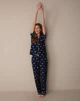 A woman wearing a navy blue pajama set with a pink and white dog print, standing with her arms raised against a plain beige studio background.