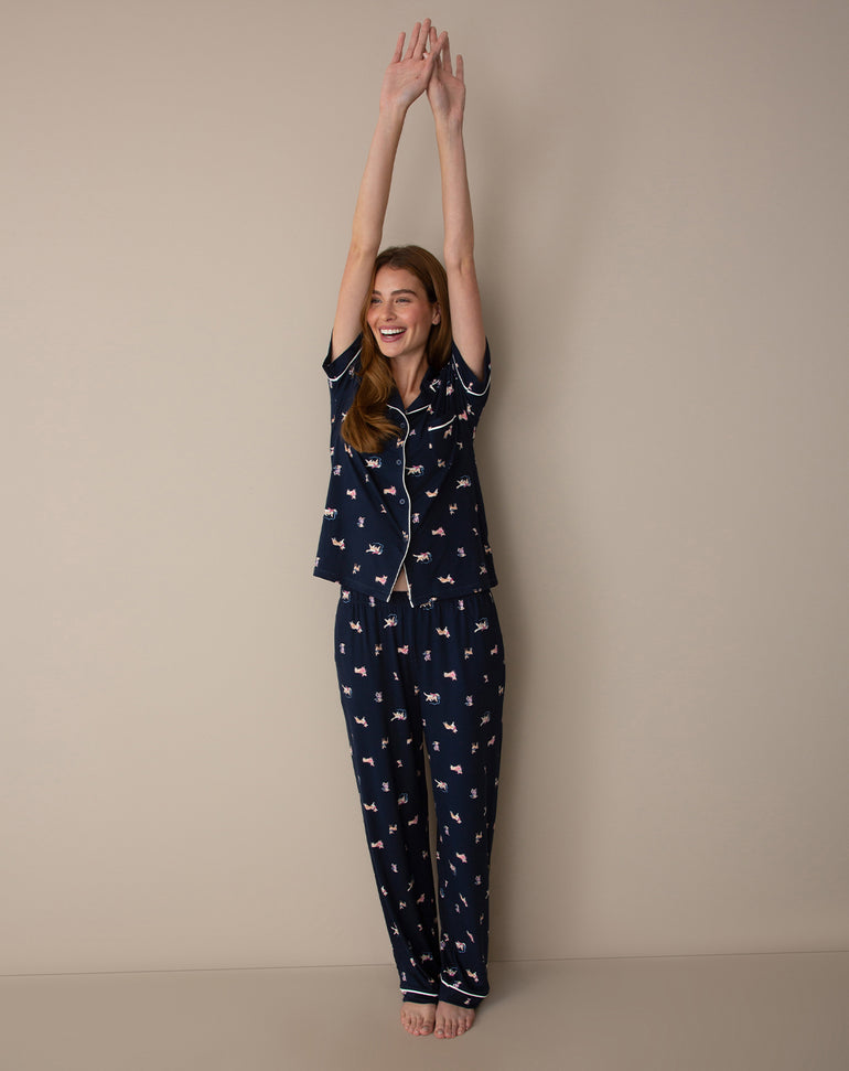 A woman wearing a navy blue pajama set with a pink and white dog print, standing with her arms raised against a plain beige studio background.