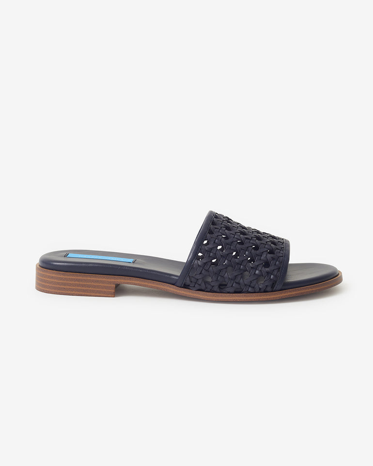 The Draper James Rattan Sandal is shown against a white background, featuring a woven navy blue upper, flat sole, and low wooden heel.