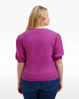 A woman with blonde hair is seen from the back wearing the Draper James Short Sleeve Cashmere Sweater in purple and blue jeans, set against a plain white background.