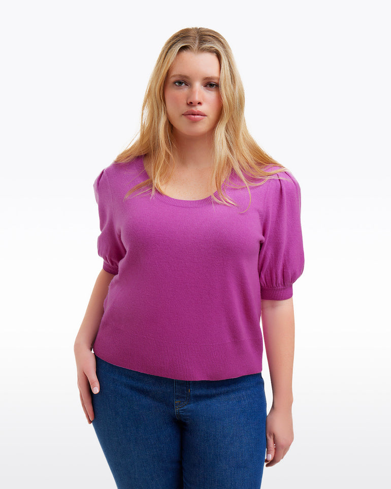 A woman with long blonde hair wears a purple Draper James Short Sleeve Cashmere Sweater and blue jeans, standing against a plain white background.