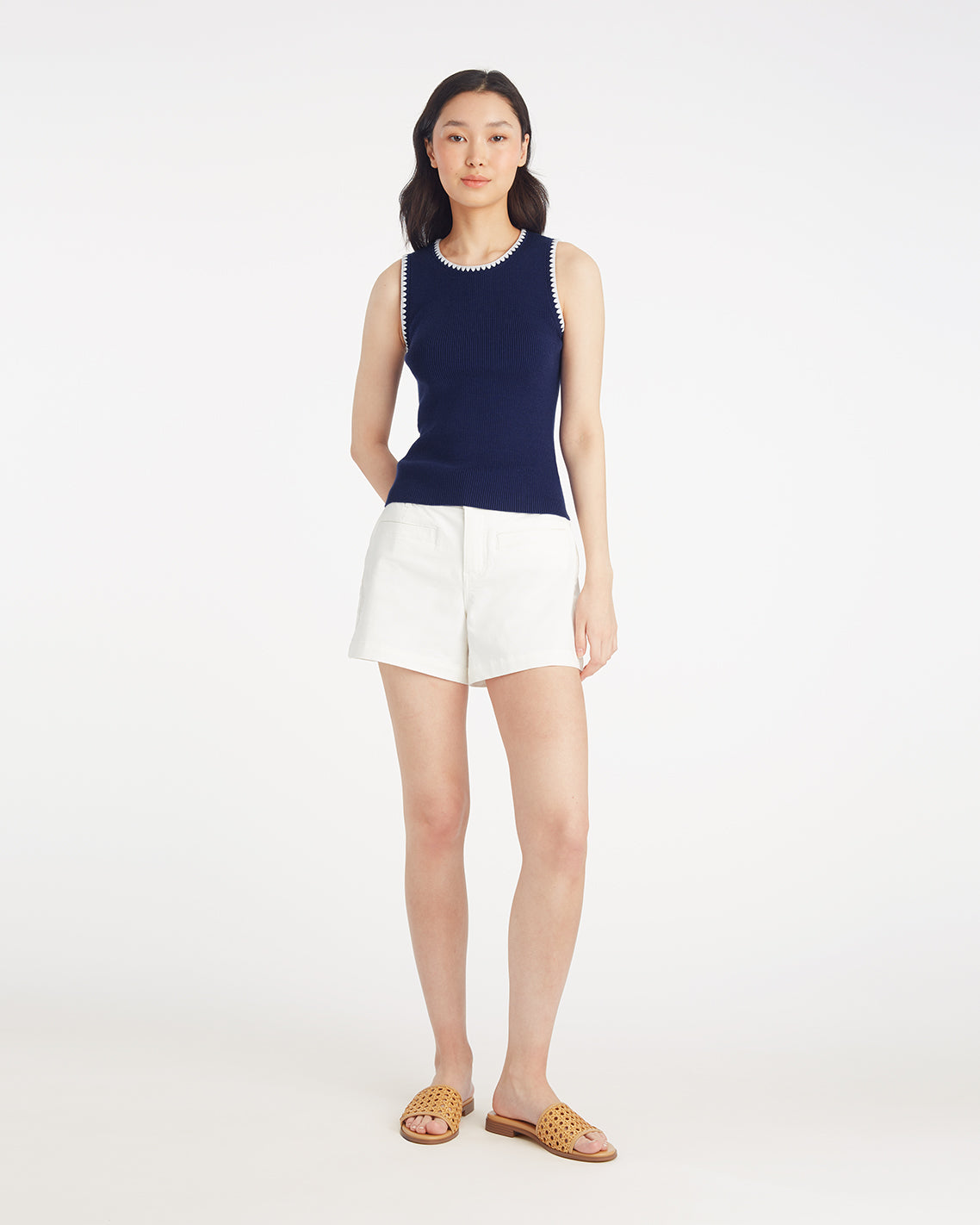 A woman stands against a plain white background wearing the Draper James Sweater Tank in navy with white trim, paired with white shorts and tan woven slide sandals.