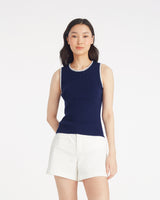 A woman stands against a plain background wearing the Draper James Sweater Tank in navy with white trim, paired with white shorts.