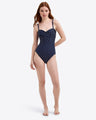 A woman poses barefoot in the Draper James Polka Dot Bandeau One Piece, a navy blue swimsuit with white polka dots and adjustable straps, ideal for a stylish beach getaway, against a plain background.