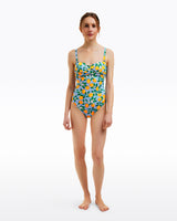 A woman faces forward wearing the Draper James Lemon Blossom Bandeau One Piece, a swimsuit with a vibrant fruit print on a light blue background.