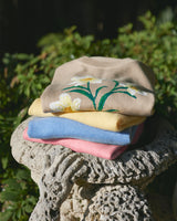 A neatly folded stack of sweaters, topped with the Draper James Daffodil Crewneck Sweater, sits on an ornate stone surface with greenery in the background.